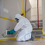 Individual in full protective gear cleaning mold off a wall.